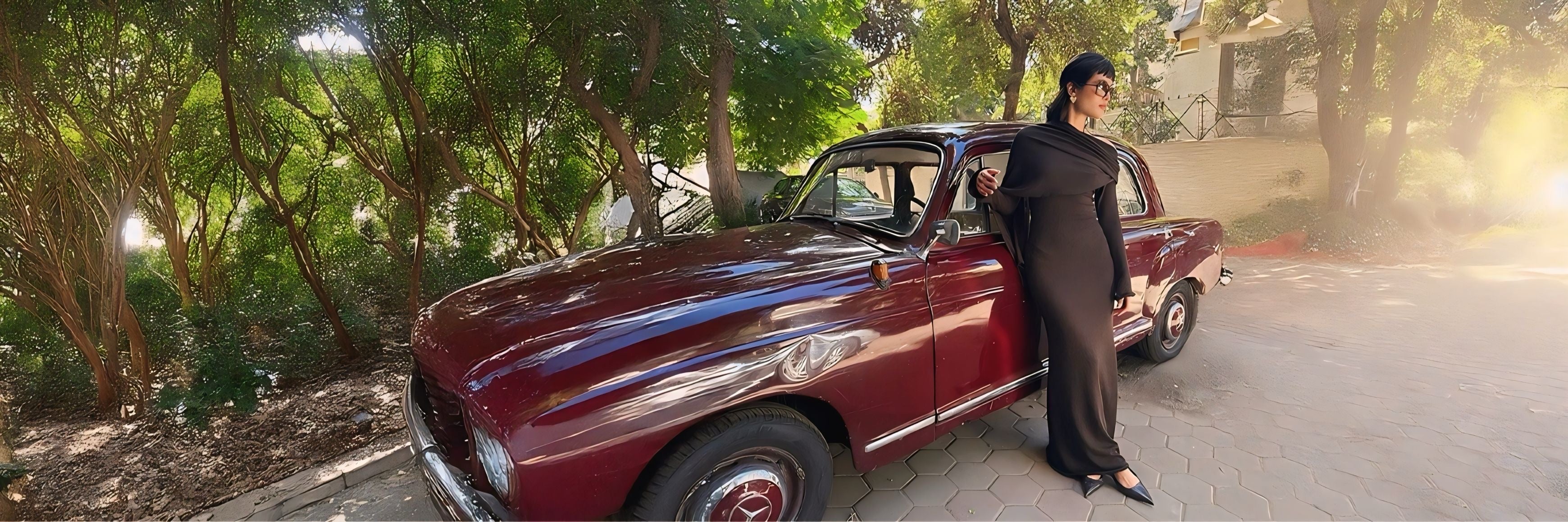 LUCE Woman in a long brown dress standing next to a vintage mercedes car on a tree-lined street.