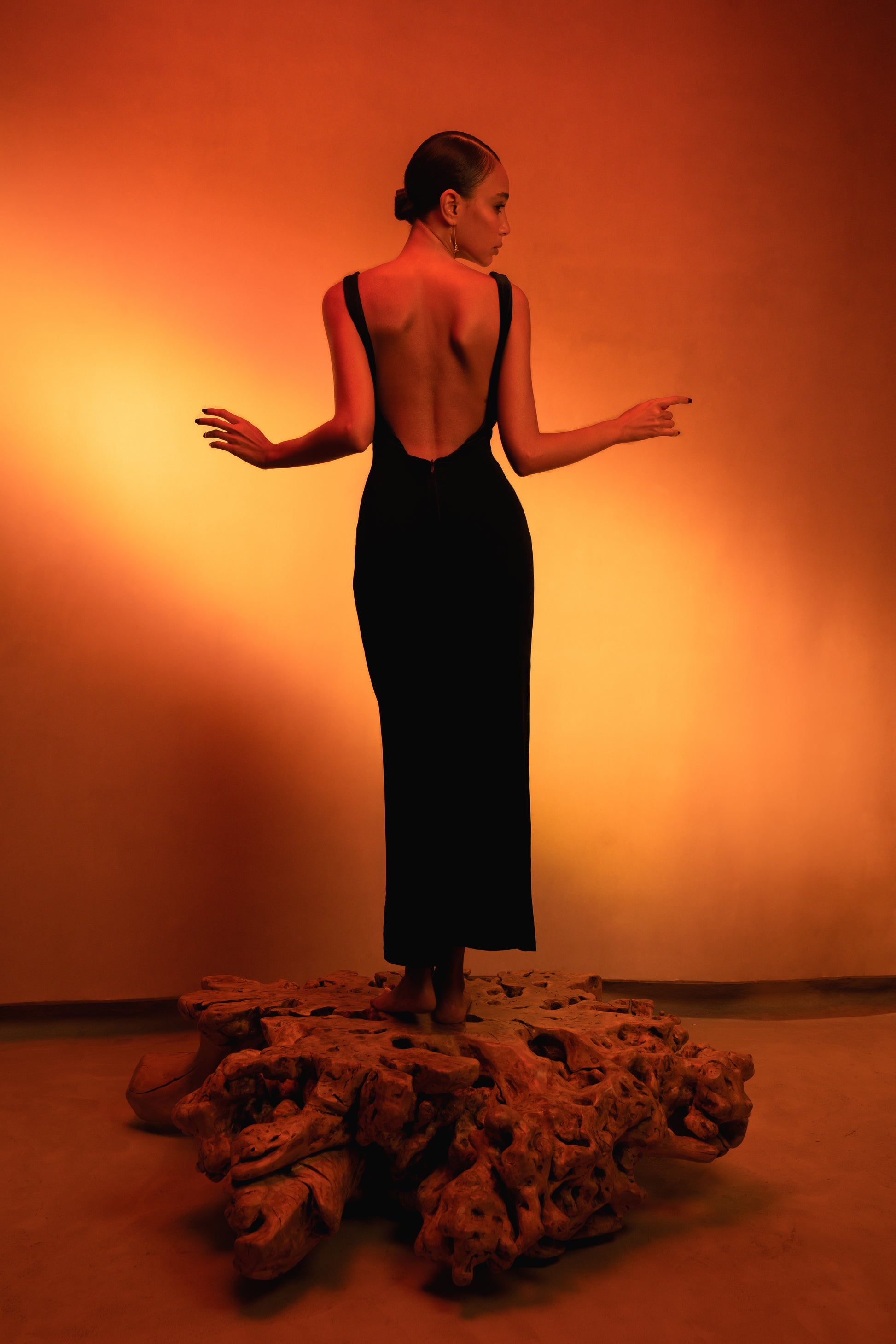 LUCE Woman in a velvet black dress with an open back standing on an old olive oil tree trunk with a warm orange background